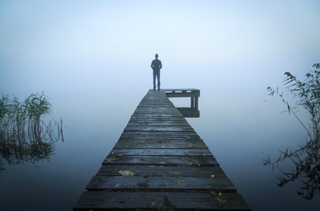 Man standing on a jetty at a lake during a foggy, gray morning.