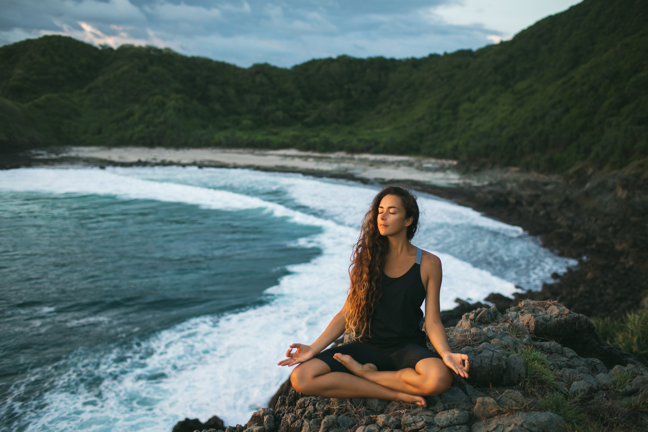 Young woman practicing yoga in lotus pose at sunset with beautiful ocean and mountain view. Sensitivity to nature. Self-analysis and soul-searching. Spiritual and emotional concept.