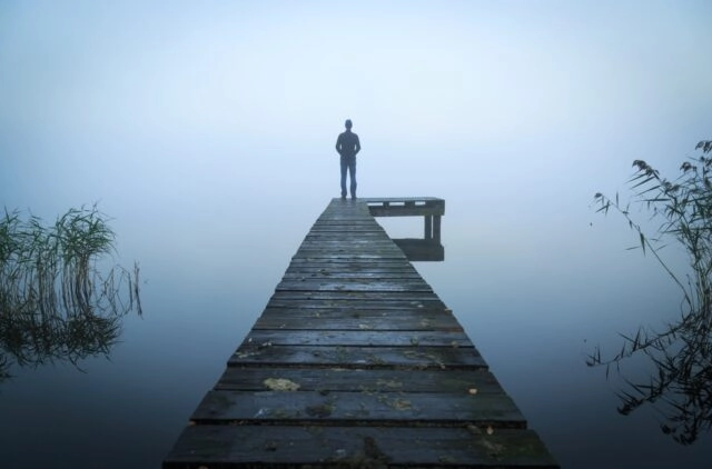 Man standing on a jetty at a lake during a foggy, gray morning.