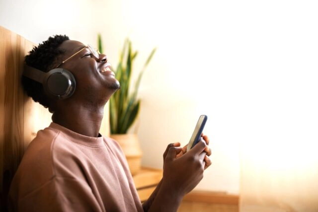 Young black man Listening to music with wireless headphones using mobile phone at home. Blissful mom Enjoyment. Young black man Listening to music with wireless headphones using mobile phone at home. Blissful moments. Copy space. Lifestyle.